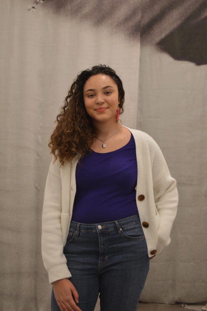Photograph of Abbi for the 21st Century Kids Project. She is standing inside the Arnolfini, Bristol. The background appears to be a black and white curtain. She is standing and smiling. She is wearing a purple top, white cardigan and blue jeans. 