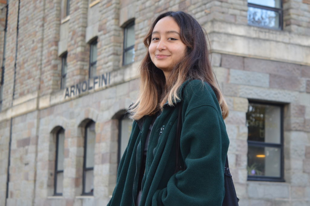 Photograph of Beatriz for the 21st Century Kid project. She is standing outside the Arnolfini in Bristol. She is looking into the camera and smiling. She is wearing a forest green fleece jacket and has a black tote bag over her shoulder.