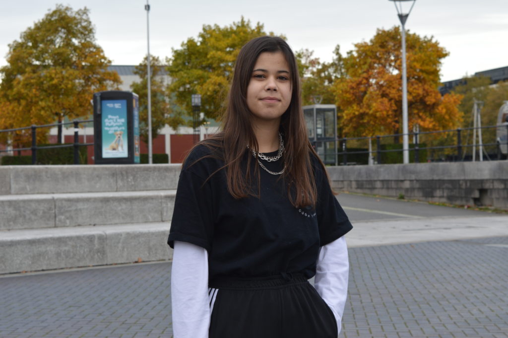 A photograph of Brooke at Millennium Square in Bristol for the 21st Century Kids project.. She is looking into the camera and wearing a black tea shirt with a white long sleeve shirt underneath and also black joggers.