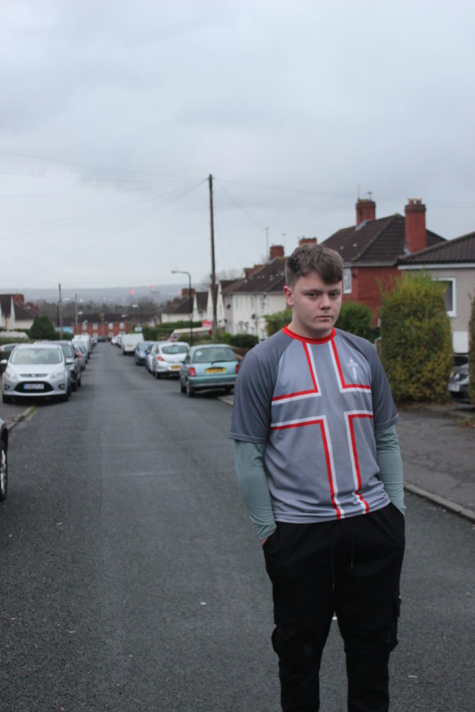 Photograph of Damien for the 21st Century Kids project. He is standing in the road in Lockleaze, Bristol. There are cars parked either side of the road and houses in the background. Damien is wearing a grey top with the St Georges cross on it and black jogging trousers