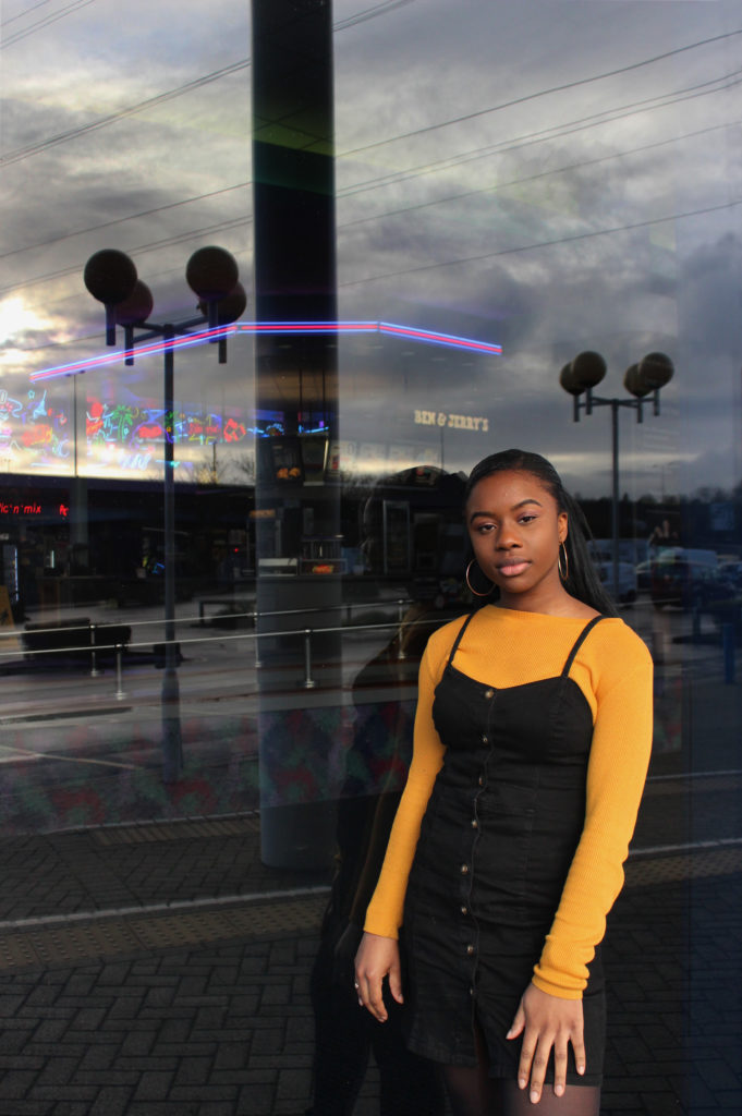 Photograph of Lydia for the 21st Century Kids project. She is beside the Showcase cinema in Avonmeads, Bristol. She is standing in front of a window. The food counter inside the cinema is just visible and the car park is reflected. She is wearing a yellow/ mustard top and black dress. 