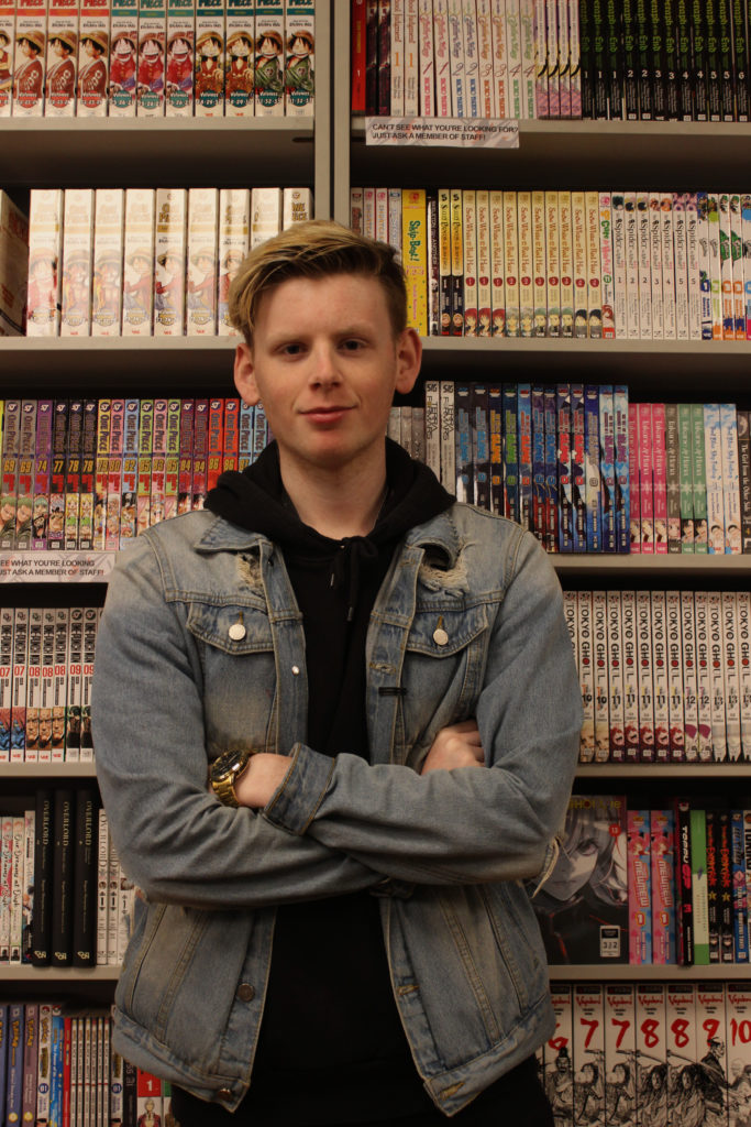 Photograph of Fred for the 21st Century Kids Project. He is standing in front of bookshelves filled with books. He is standing with his arms crossed and smiling in to the camera. He is wearing a black hoodie and a denim jacket.