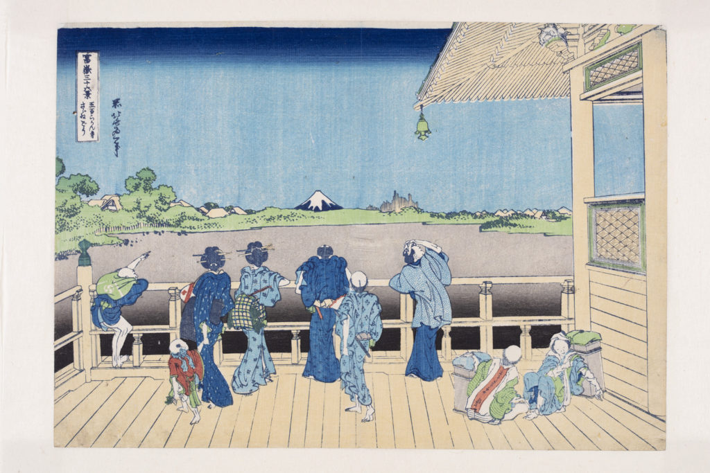 Japanese print of a group of people in elaborate traditional robes standing on a veranda looking out over the landscape. One of the porters points out Mount Fuji in the distance to them. Other porters sit on the floor resting the heavy boxes.