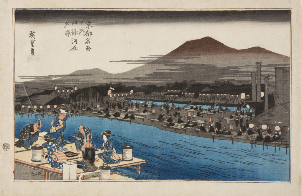 Japanese print of a landscape, in the foreground a group of two men and three women (geisha), dressed in traditional clothes, sit on a platform eating by the river bank, one woman stands and holds a fan to her face. On the other side of the river there are lots of other platforms and people, mountains rise up behind.