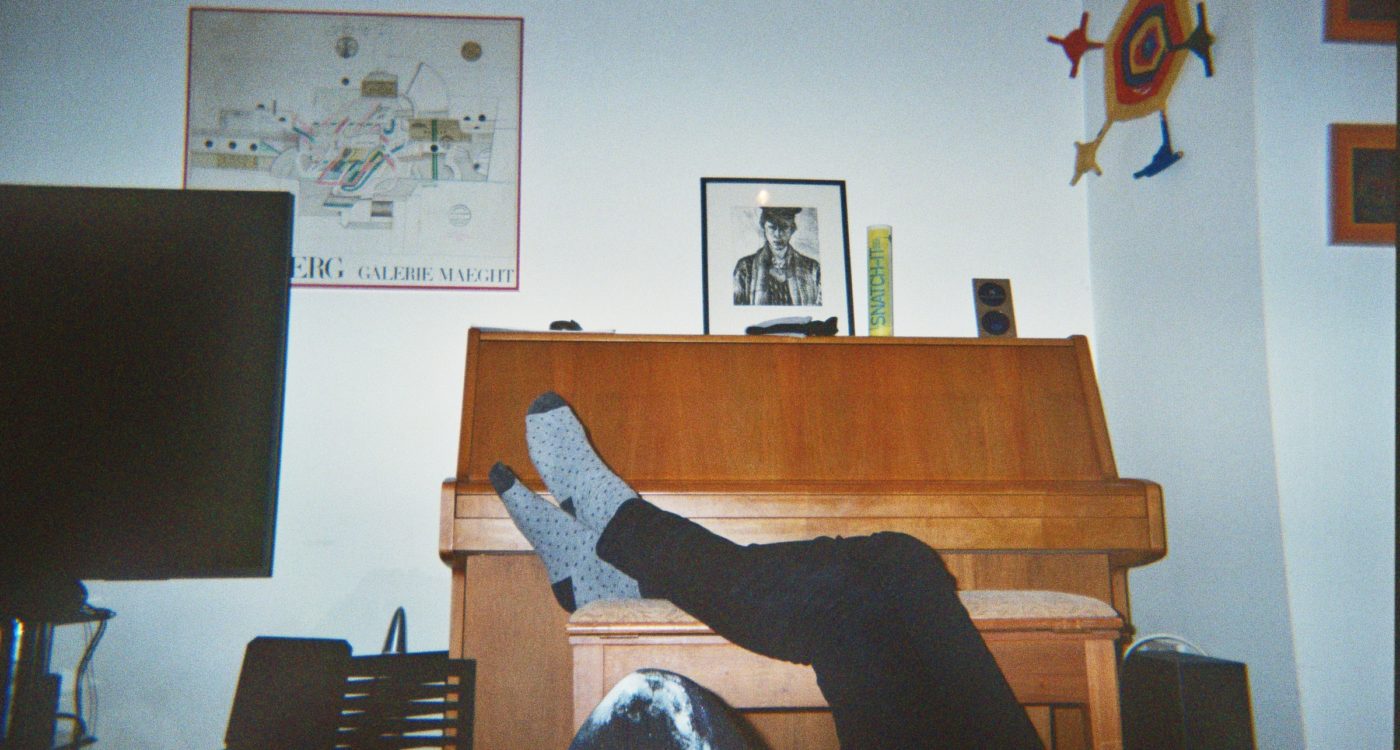 Elio's photograph (for the 21st Century Kids) of a room with a TV, pictures on the wall, and a piano. Someones legs are resting on the piano bench.