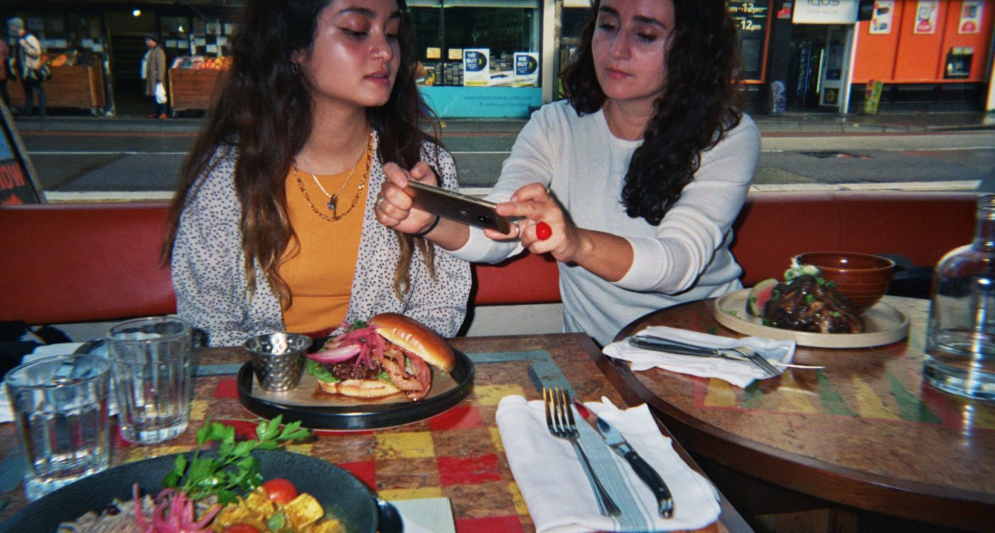 Beatriz's photograph (for the 21st Century Kids) of her sister and her mum at the restaurant Turtle Bay. They are sitting at the table with their meal and her mum is taking a picture of the burger whilst her sister watches her.