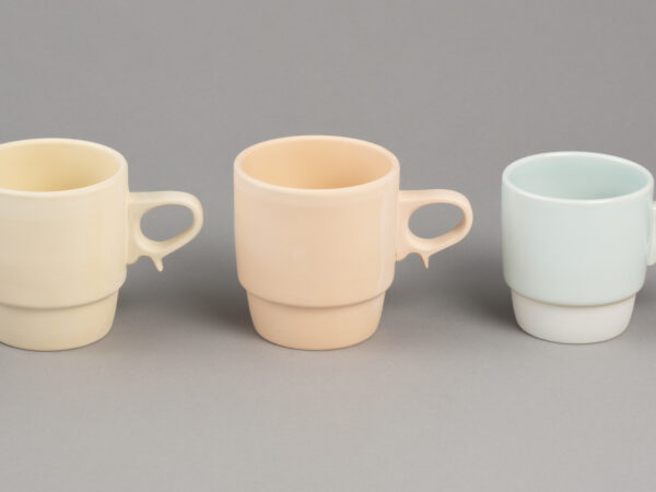 Three porcelain mugs in different stages of production, with yellow, pink and blue colour tones