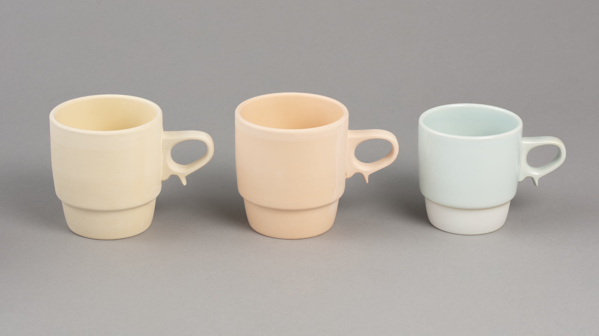 Three porcelain mugs in different stages of production, with yellow, pink and blue colour tones