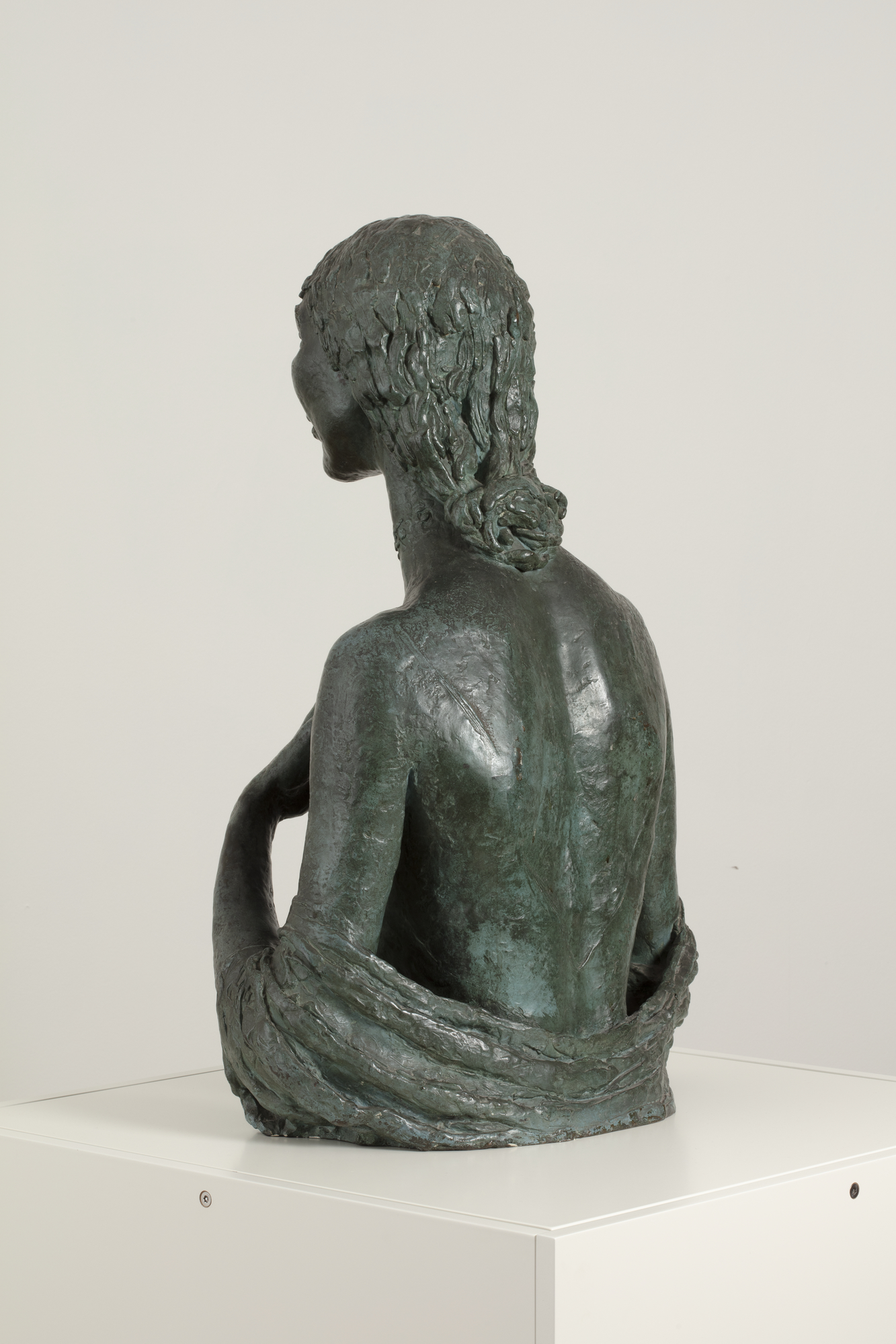 A rear view of a bronze sculpture of a naked woman shown from the waist up, showing an elaborate knotted hair style.