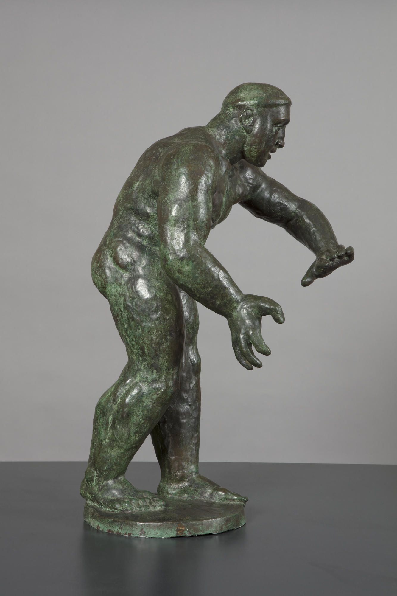 A bronze sculpture of a naked man shown as well-muscled wrestler facing right, with thick legs and arms, who leans inwards with hands poised as if to grapple an opponent.