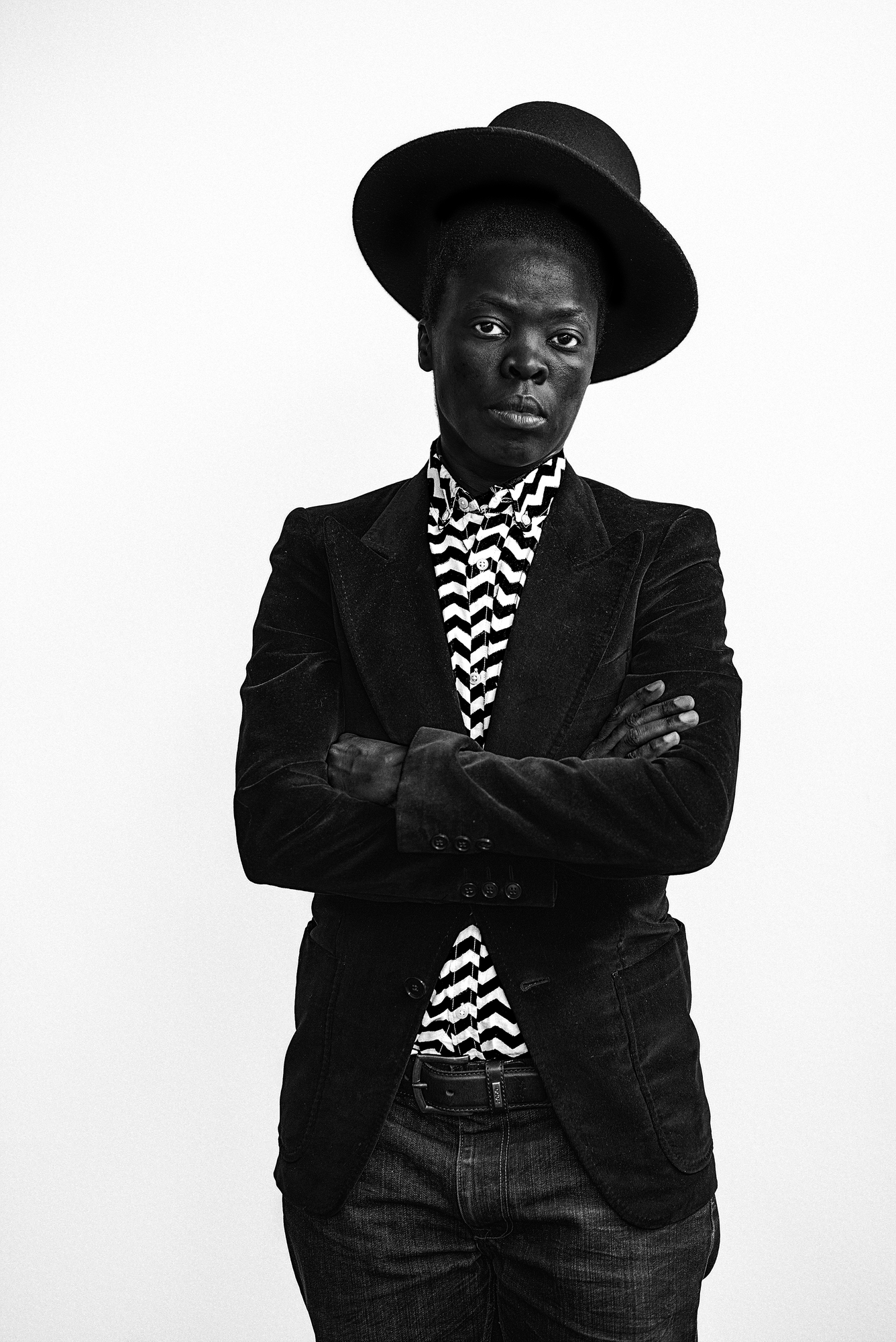 A black and white photographic self-portrait Zanele Muholi from the hips upwards with arms folded across the chest, shown wearing a hat at a jaunty angle, jeans, a black velvet jacket, and a black and white zig-zagged patterned shirt.
