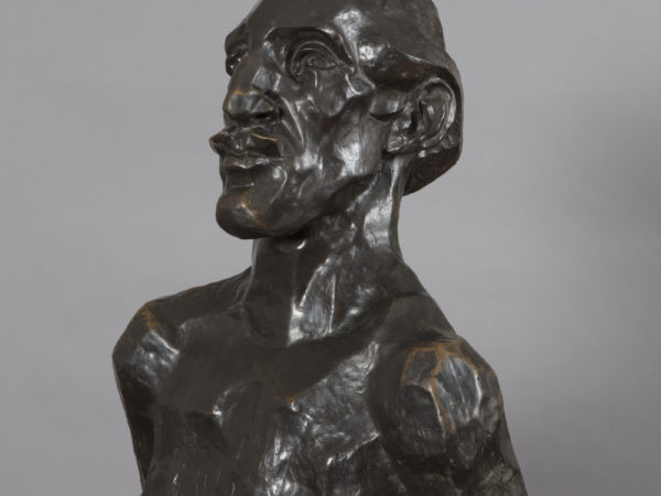 A bronze sculpture of a naked man's head and shoulders, viewed facing half-right , with an angular face and prounced chin, nose and lips as well as a short swept back receding hair style. His bare chest has been scratched to represent a tattooed design.