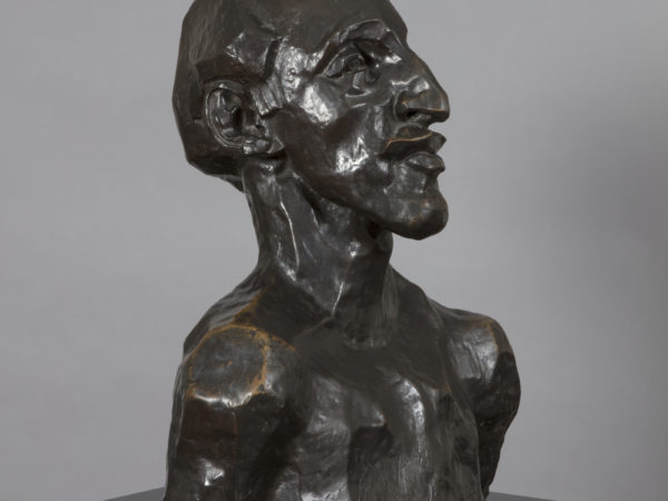 A bronze sculpture of a naked man's head and shoulders, viewed facing half-left, with an angular face and prounced chin, nose and lips as well as a short swept back receding hair style. His bare chest has been scratched to represent a tattooed design.