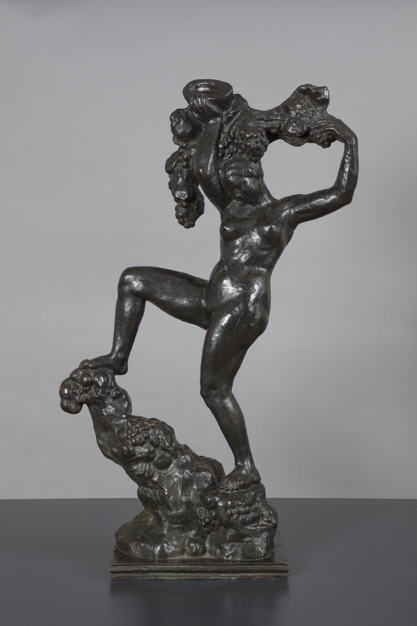A bronze sculpture of a naked woman balancing on a rocky peak with her right leg held high and bent at the knee. Her arms are raised above her head holding a vine with bunches of grapes.