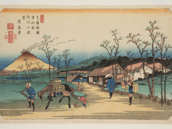 Japanese print of a road, leading to a village and in the distance are mountains. In the foreground are travelers. A man leads a horse carrying boxes, a small figure follows behind scooping up horse manure. A man walks towards the village with an attendant, carrying parcels, following behind.