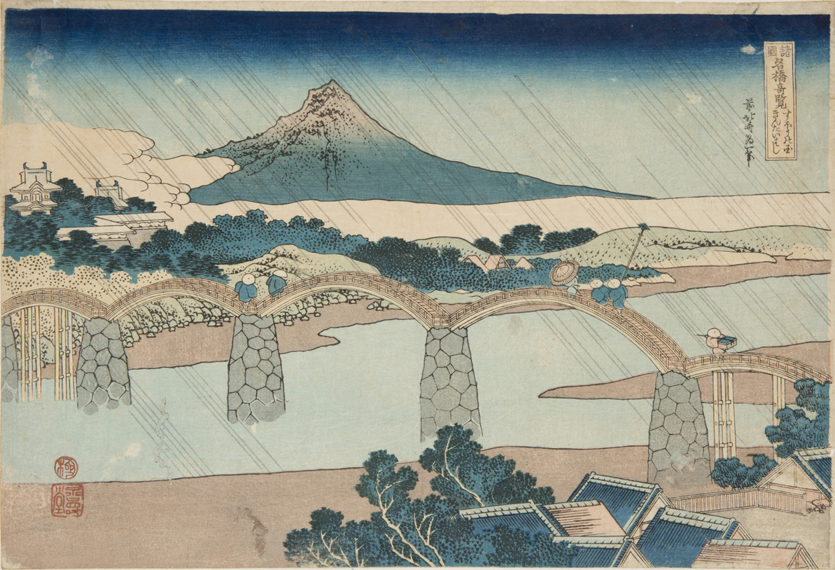 Japanese print of a landscape. Rain slants across the scene top left to bottom right. In the foreground there is a river bank with trees and roof tops. Middle is a bridge spanning the river with five arches and people standing on it. In the background there are a few buildings, trees, fields and an imposing mountain.