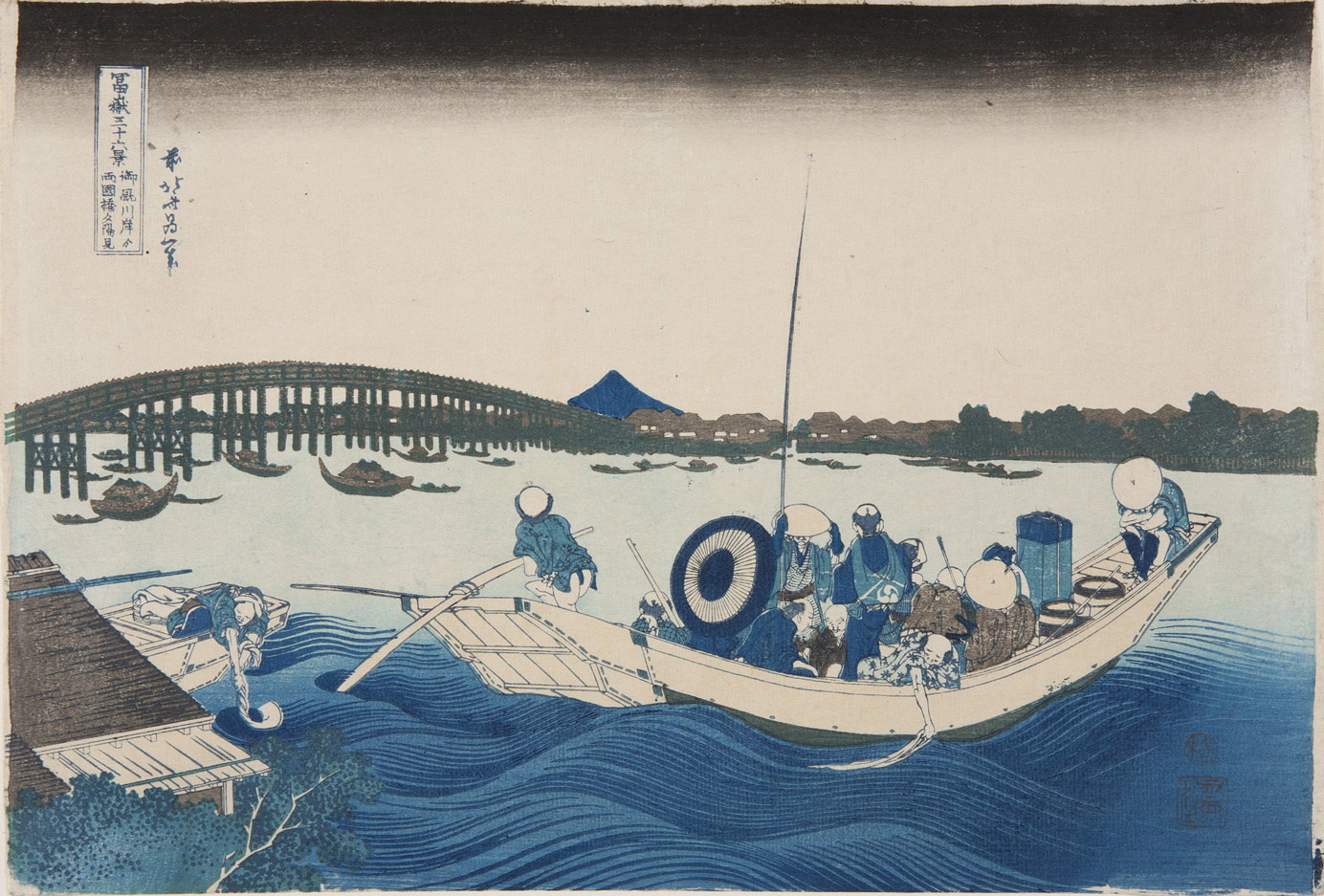 Japanese print of a river scene. In the foreground there are waves and a boat full of passengers, the boatman steers with a pole. Many boats are on the water and a bridge spans the river. The far bank has buildings and trees and Mount Fuji is in the distance.