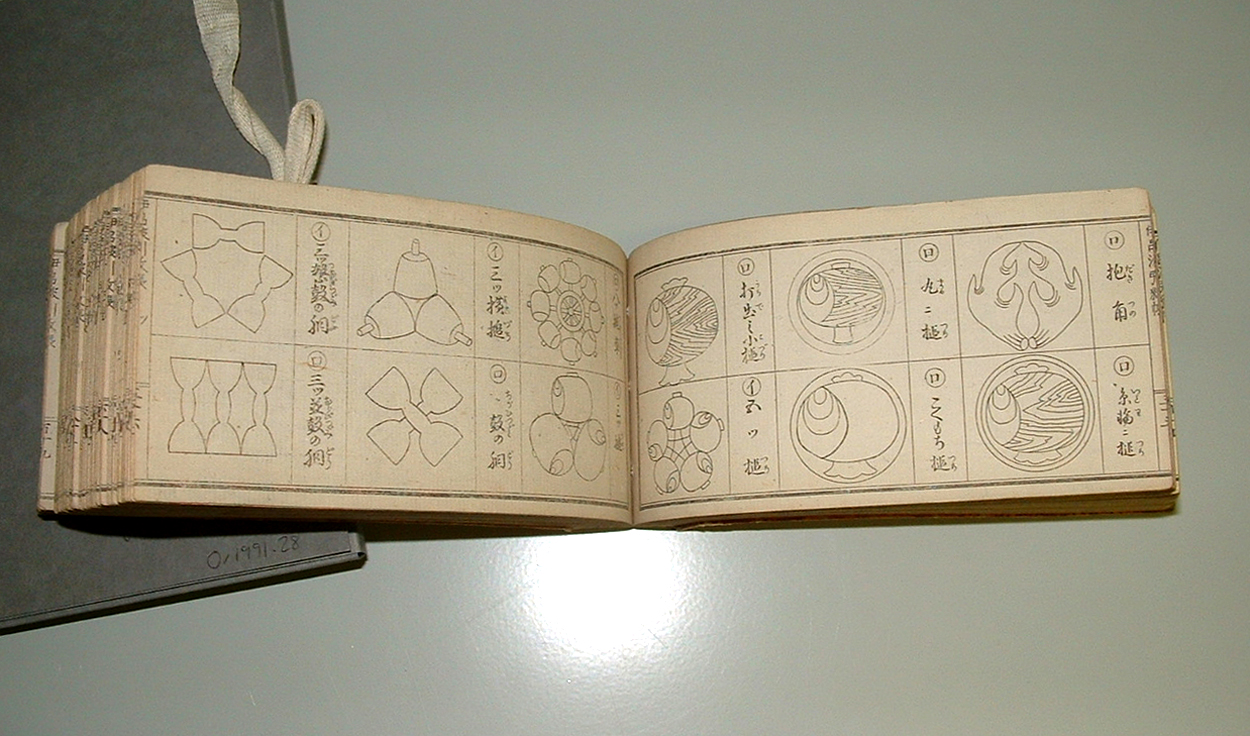 An open book shows line drawings of Japanese family crests, six on each page, and Japanese text.