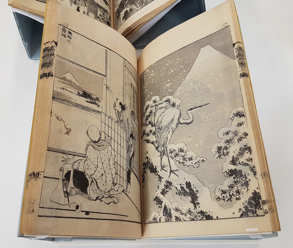 Image of an open Japanese book showing two pictures. One of two figures dressed in traditional costume looking out of a window at Mount Fuji. the other of a standing stork in a snowy landscape with Mount Fuji behind.