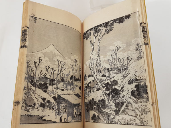 Image of open pages of a Japanese book showing a landscape, in the foreground is a building with people passing by, dressed in traditional clothes walking along the road that leads over the hills, through the trees, in the background is Mount Fuji.