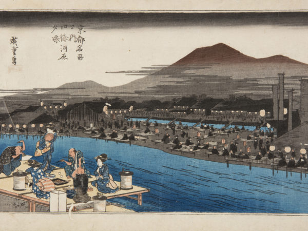 Japanese print of a landscape, in the foreground a group of two men and three women (geisha), dressed in traditional clothes, sit on a platform eating by the river bank, one woman stands and holds a fan to her face. On the other side of the river there are lots of other platforms and people, mountains rise up behind.
