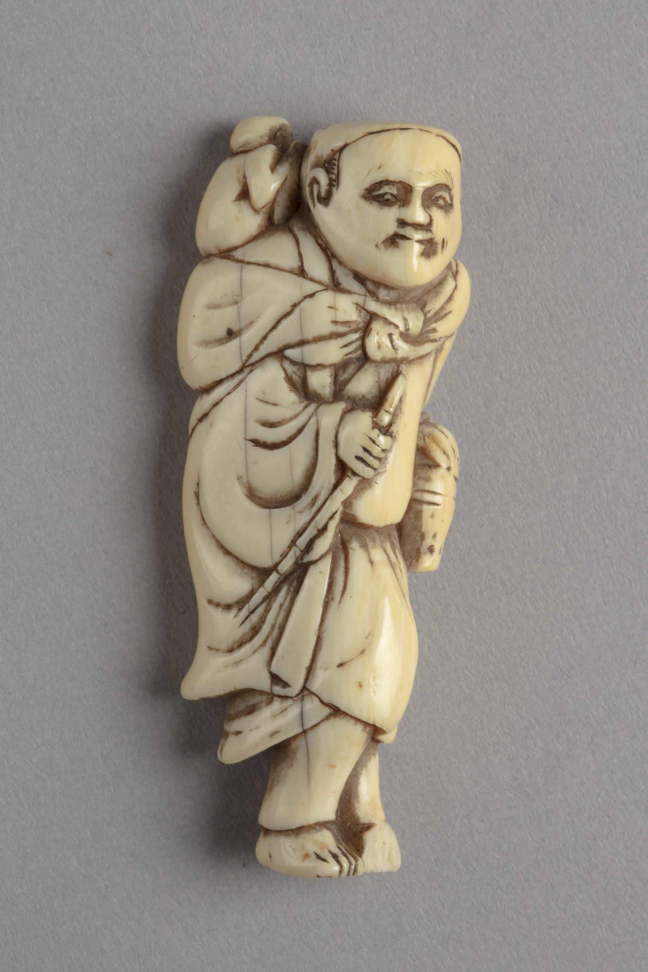 A Japanese ivory netsuke of a standing man holding a stick and alms basket with a small monkey on his shoulder.