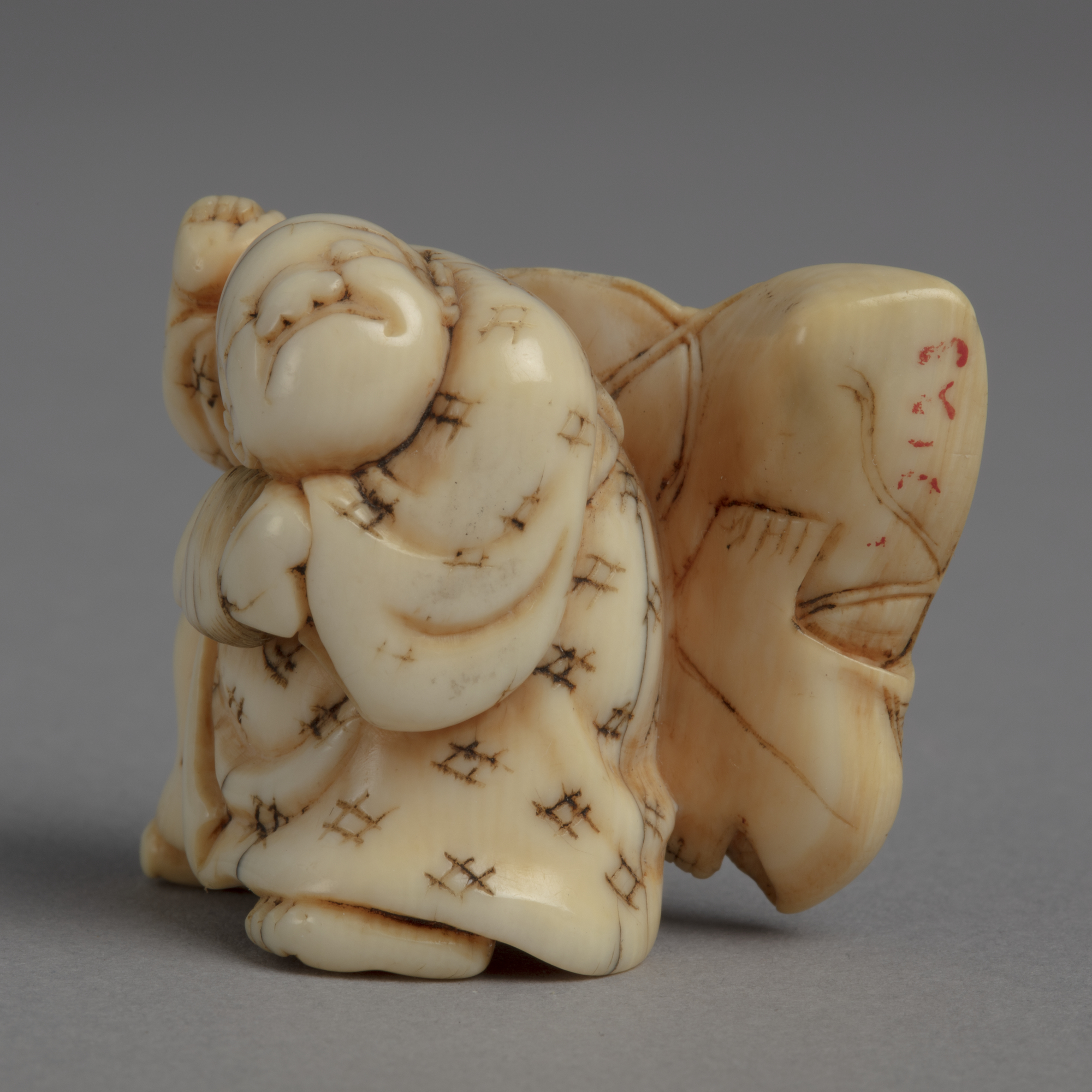 A Japanese ivory netsuke of a boy with a reel in his left hand and a kite over his back painted with a stern face.