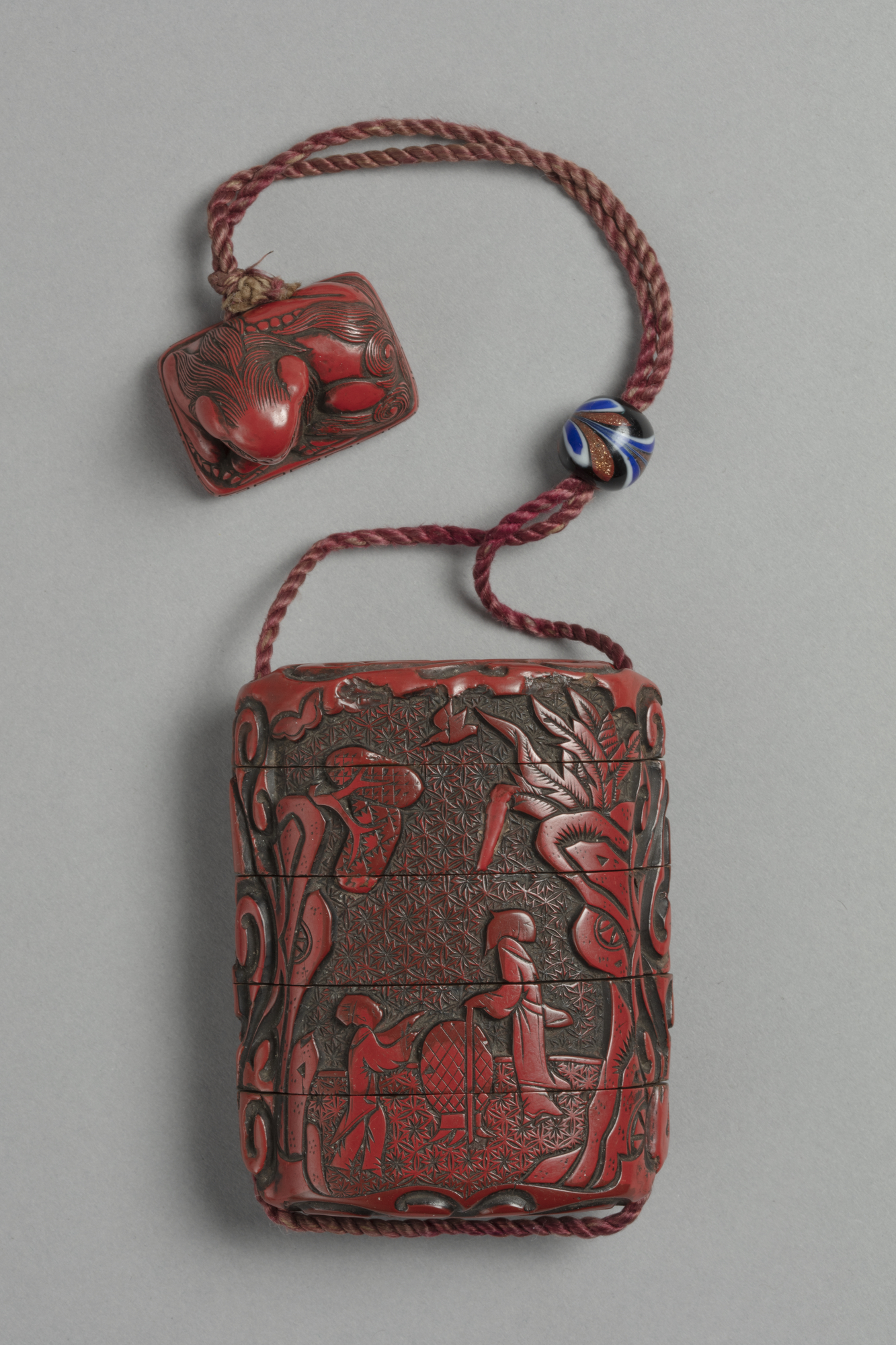 A Japanese red lacquer inro box with five compartments carved with a garden landscape, strung to a carved netsuke toggle in the form of a lion.
