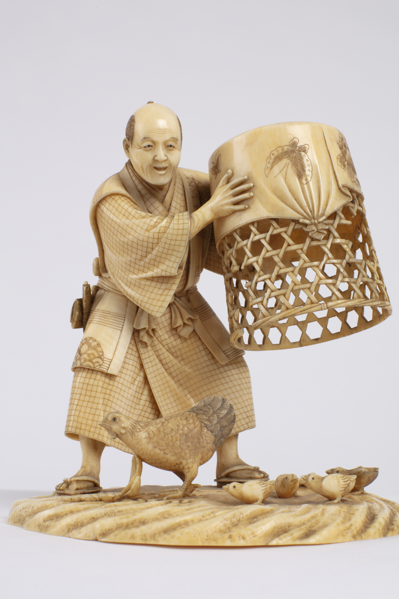 A Japanese carved ivory okimono ornament of a man with kimono, netsuke and tobacco pouch holding a chicken coop.