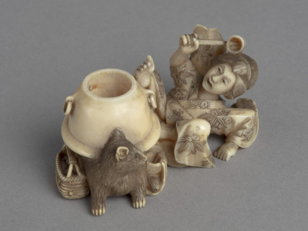 A Japanese ivory okimono ornament of a lady fallen on her back in alarm at the half-badger half-kettle before her.