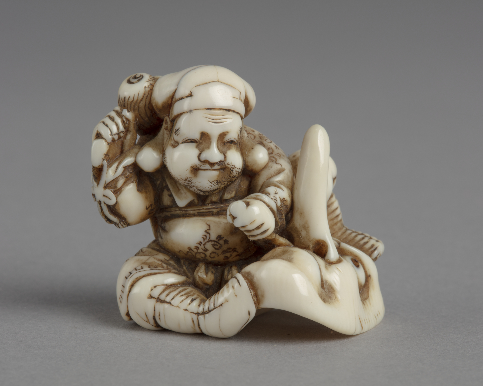 A Japanese ivory netsuke of Daikoku chiselling out the nostril of a long-nosed mask using his magic mallet.