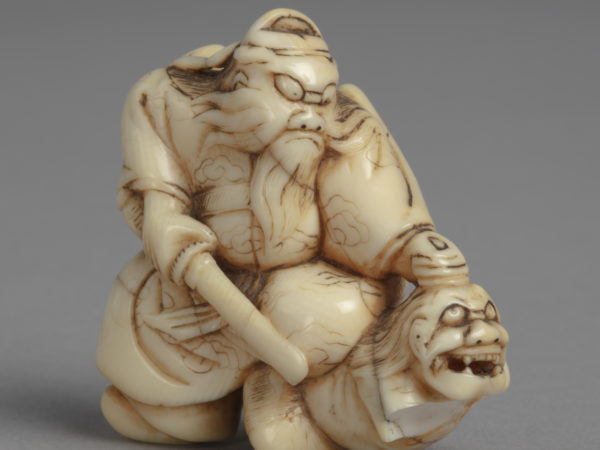 A Japanese ivory netsuke of Shoki kneeling on a demon, grasping it by the hair and pointing a sword at it.