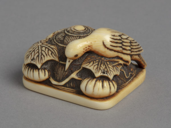 A Japanese ivory netsuke of a stork bird perched on a large straw hat discarded among pumpkins, vines and leaves.