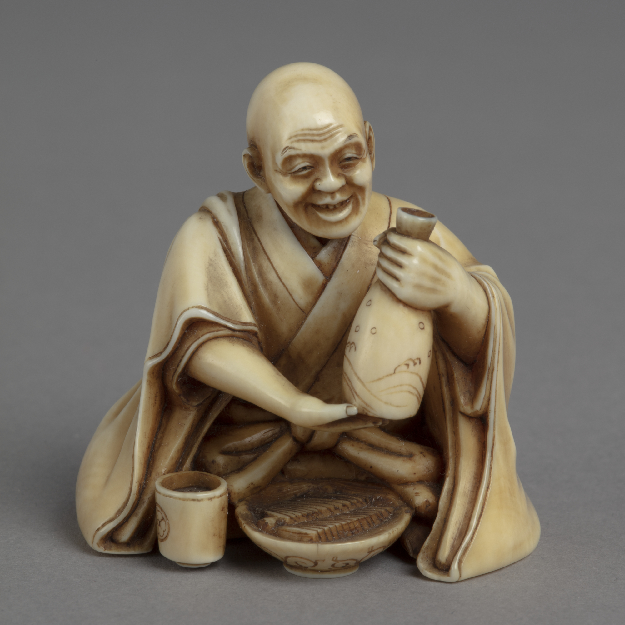 A Japanese okimono ornament of a seated man holding aloft a sake flask, a bowl of noodles and cup before him.