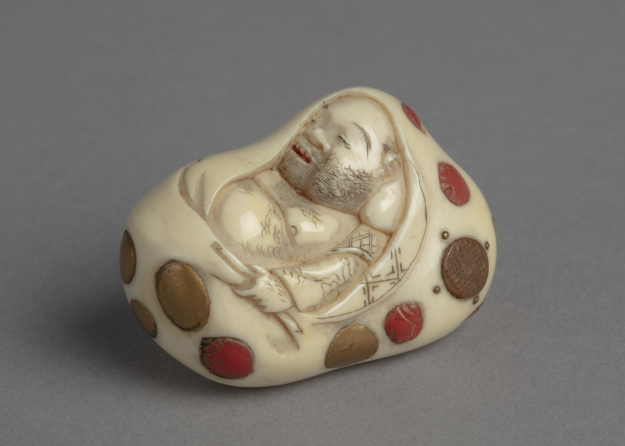 A Japanese ivory netsuke inlay with abalone, red coral, wood and gold paint, of Hotei asleep in his treasure bag.