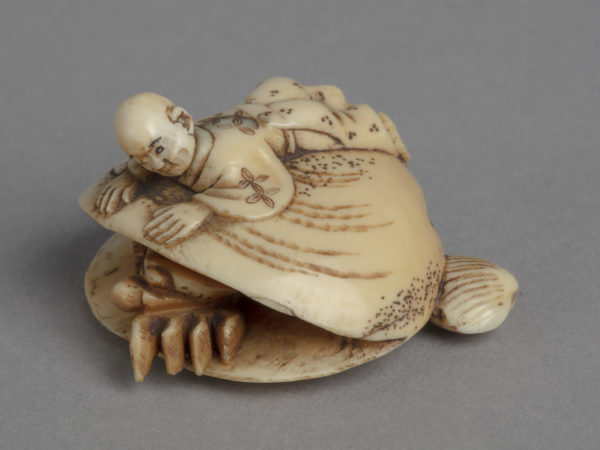 A Japanese ivory netsuke of a boy lays on his front on the back of a large clamshell, from which a crab emerges.