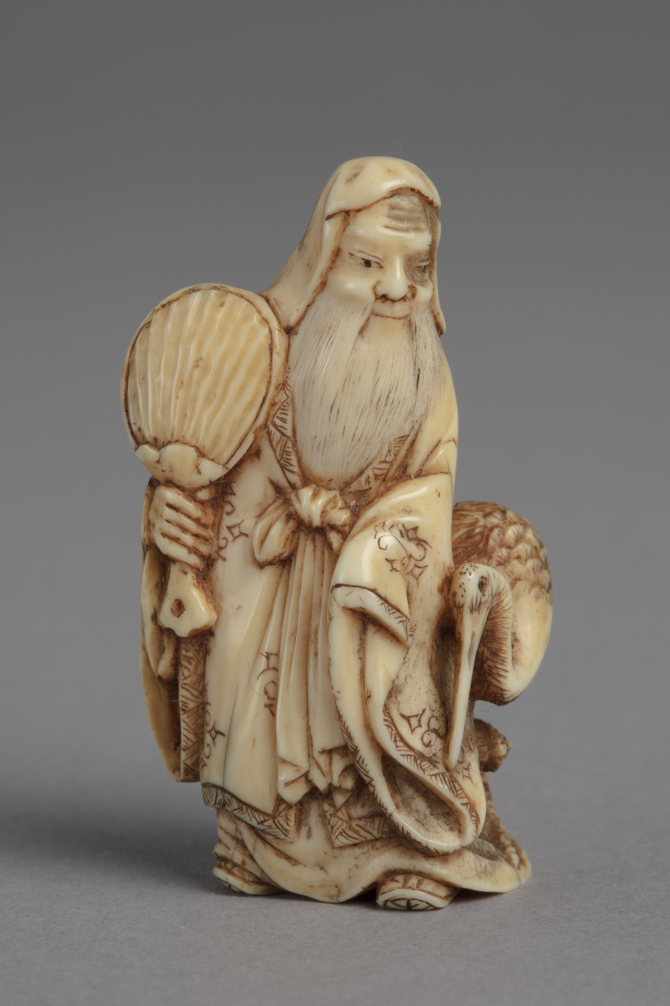 A Japanese ivory netsuke of Fukurokuju a god of wisdom. He stands holds a fan in his right hand, a stork by his left side.