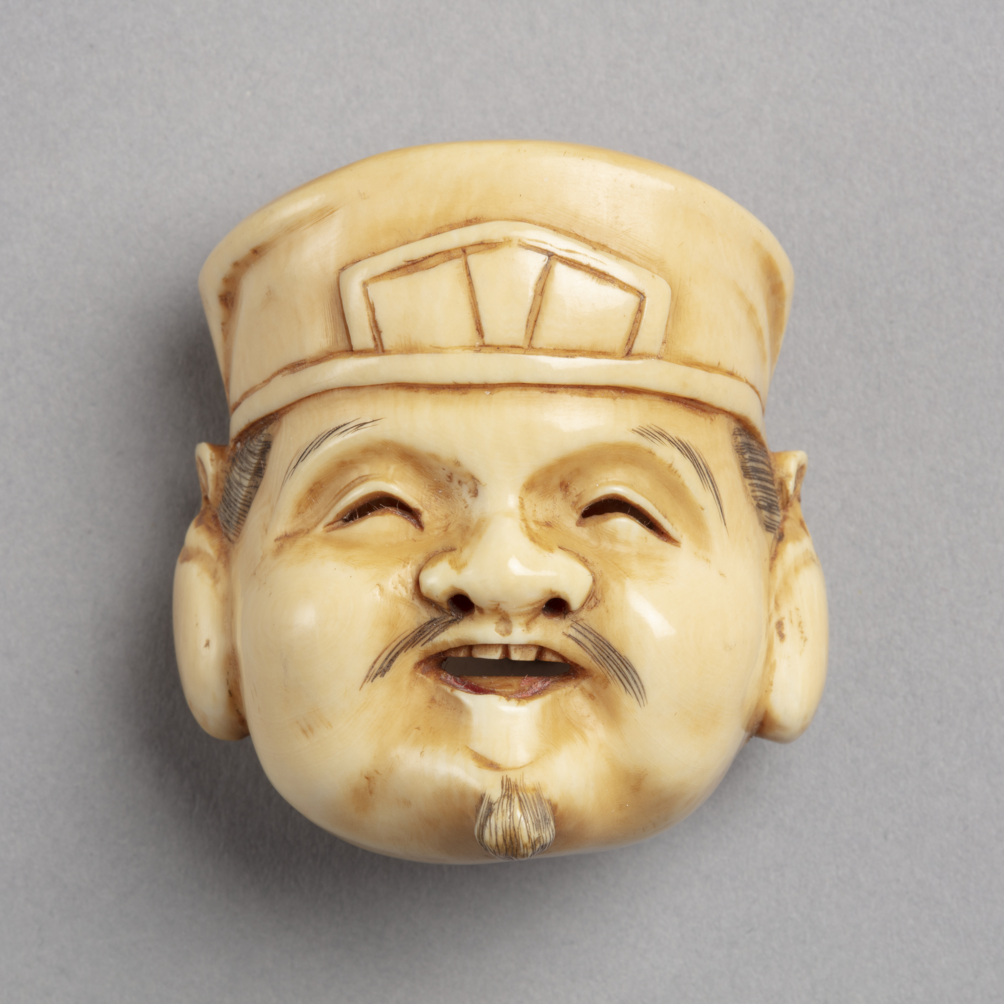 A Japanese ivory netsuke of the smiling face of Ebisu. He wears a hat and has a fine moustache and small beard.