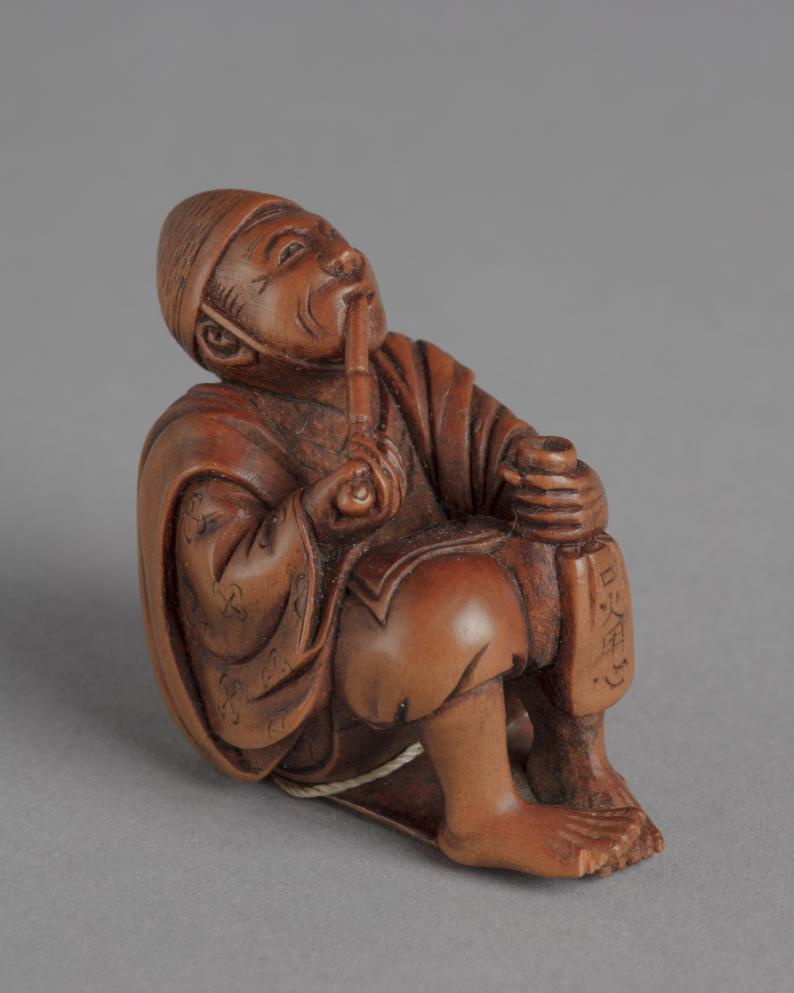 A Japanese boxwood netsuke of a seated man looking up and smoking a long pipe, left hand on left knee and holding a bag.