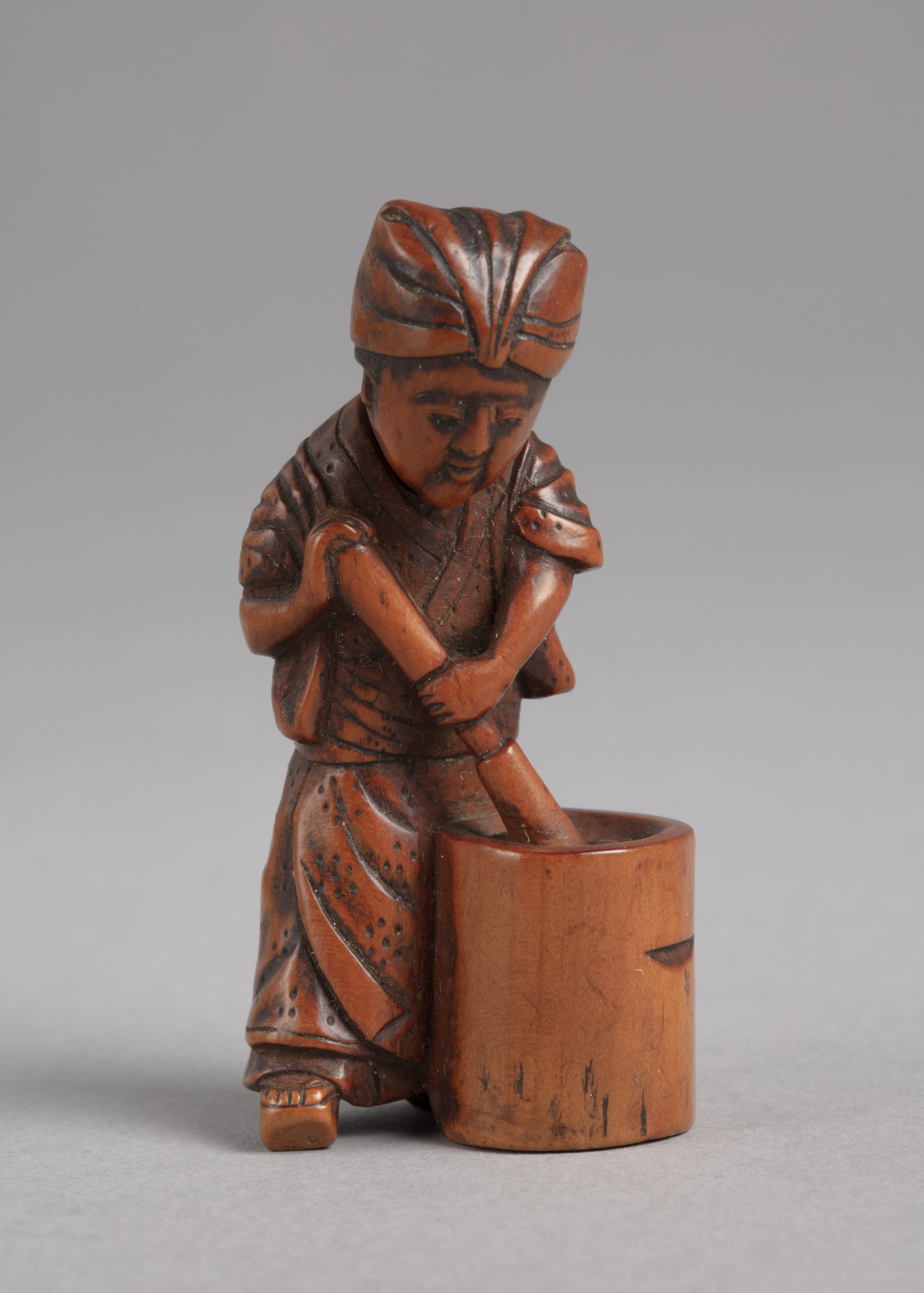 A Japanese boxwood netsuke of a standing woman with movable head washing clothes in a cylindrical tub with a pestle.