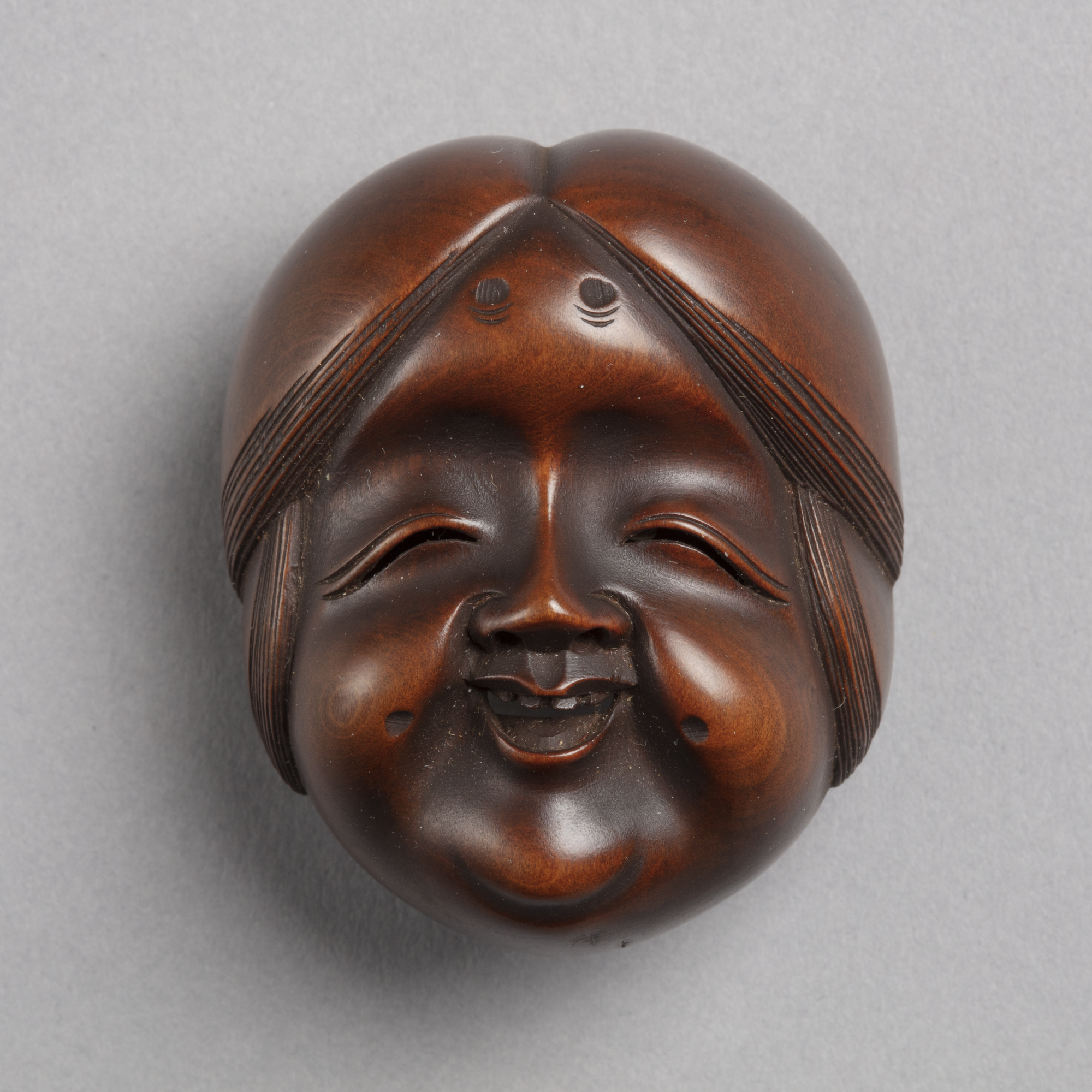A Japanese boxwood netsuke of the face of Okame, the goddess of mirth. She has large cheeks and a joyful smile.