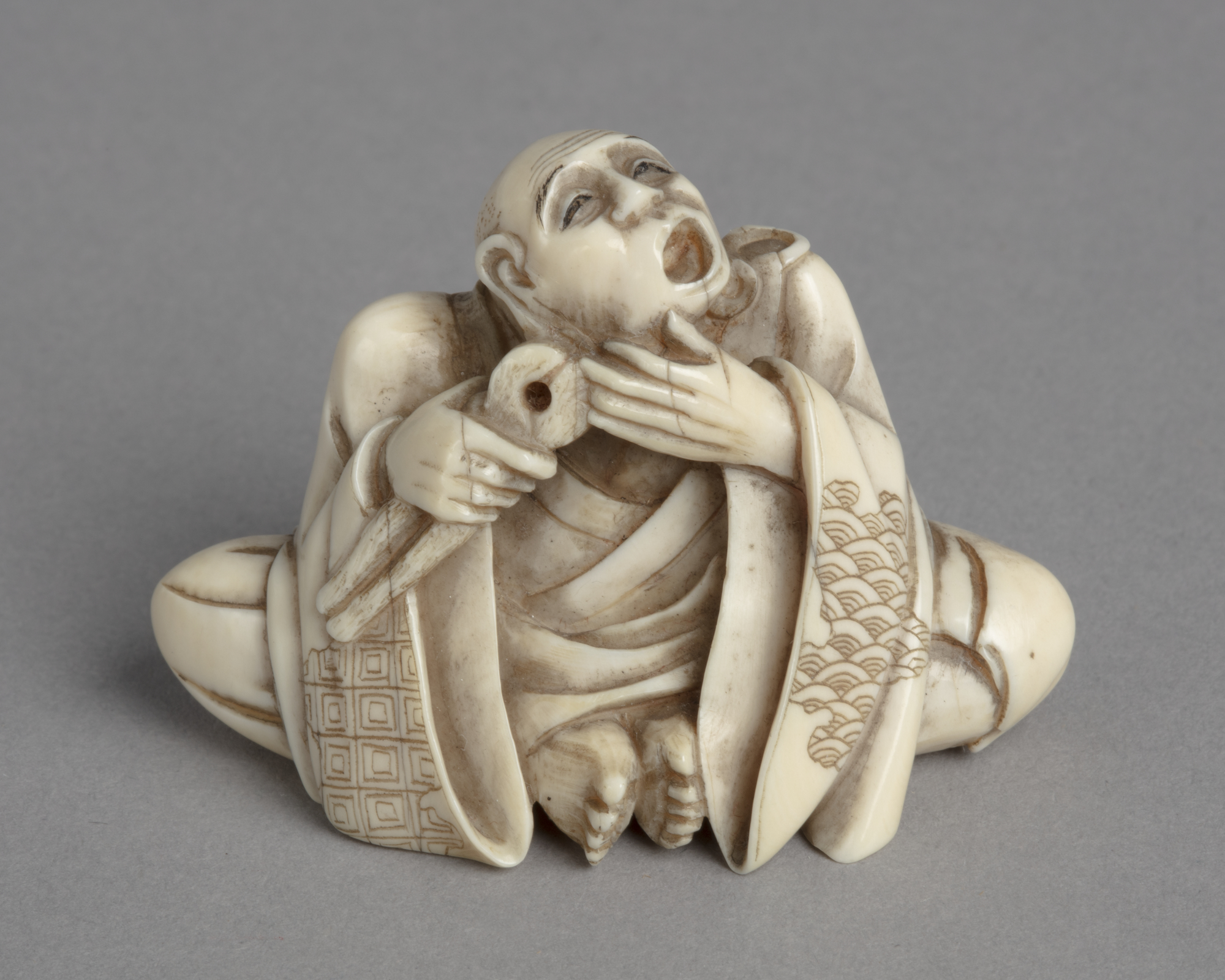 A Japanese ivory netsuke of a man in robes seated with feet together holding pliers to his mouth.