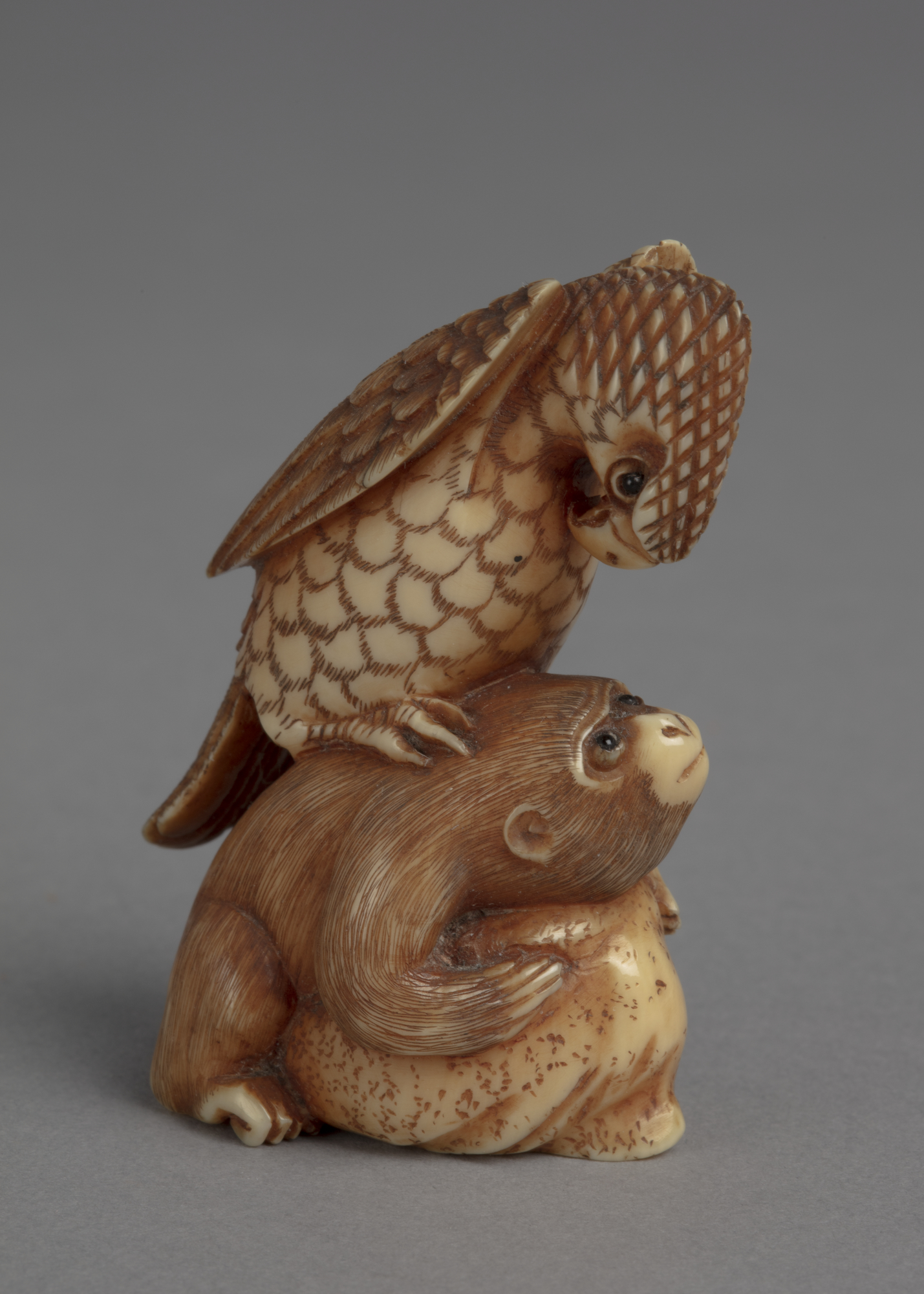 A Japanese ivory netsuke of a hawk perched on the back of a monkey that is crouching over a rock.