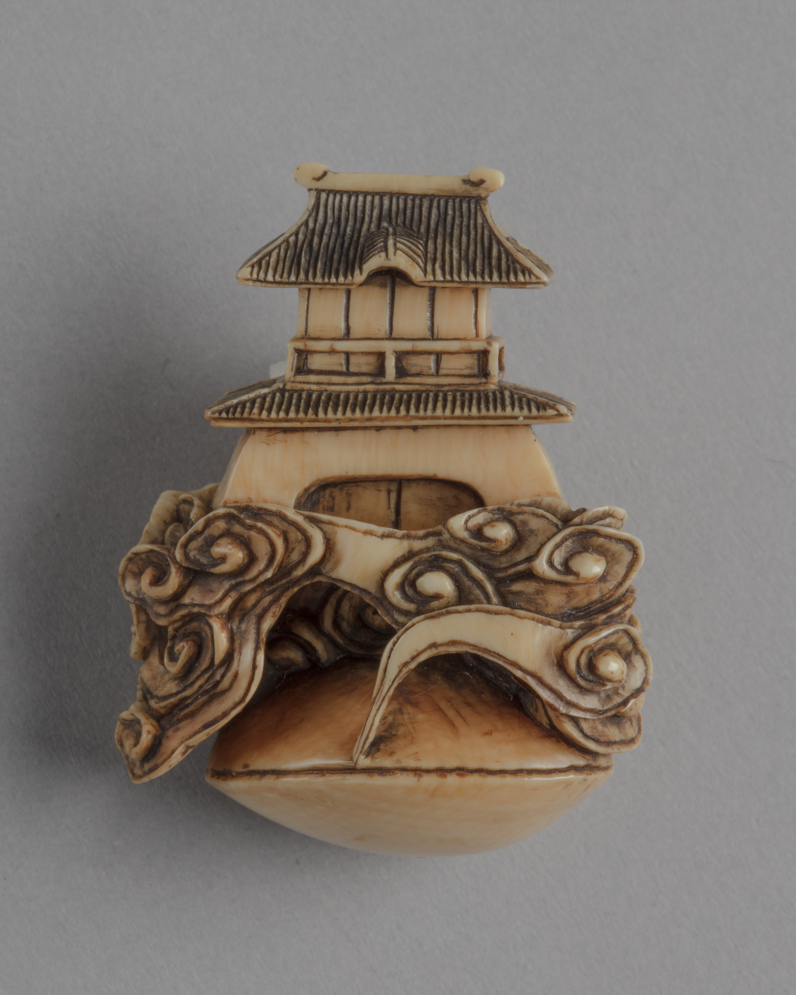 A Japanese ivory netsuke of a two-roofed gate-tower rising out of clouds emerging from a closed clam shell.