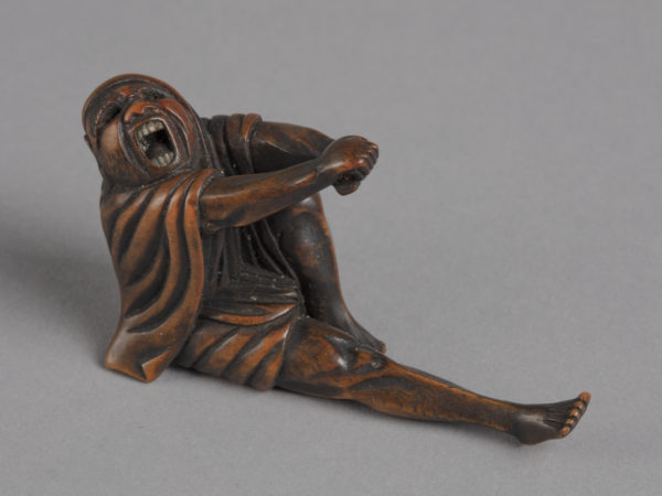 A Japanese boxwood netsuke of the monk Daruma yawning, stretching out clasped hands and right leg.