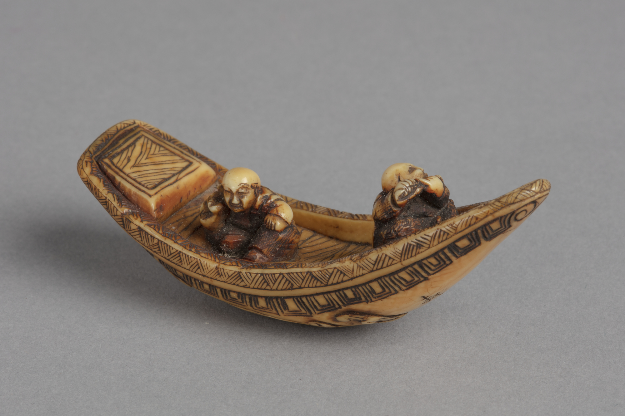 A Japanese ivory netsuke of two seated men, one playing a flute, in a long wooden boat with waves underneath.