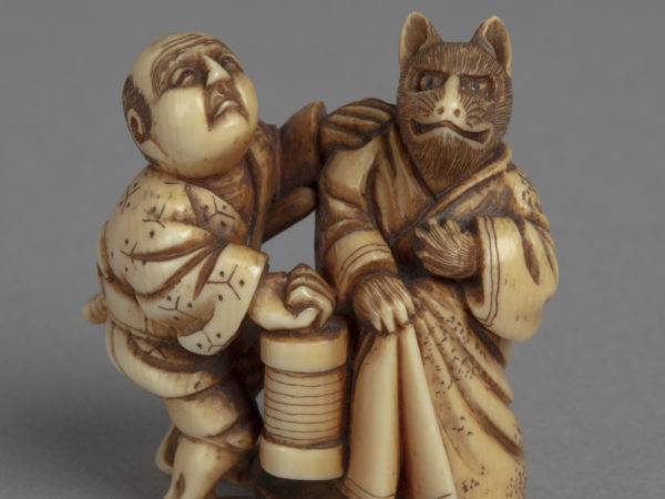 A Japanese ivory netsuke of a kitsune fox dressed in a kimono standing to the left of a man who carries a lantern.