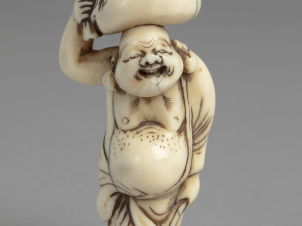 A Japanese ivory netsuke of Hotei, a god of happiness, standing with large belly exposed and carrying a bag on his head.