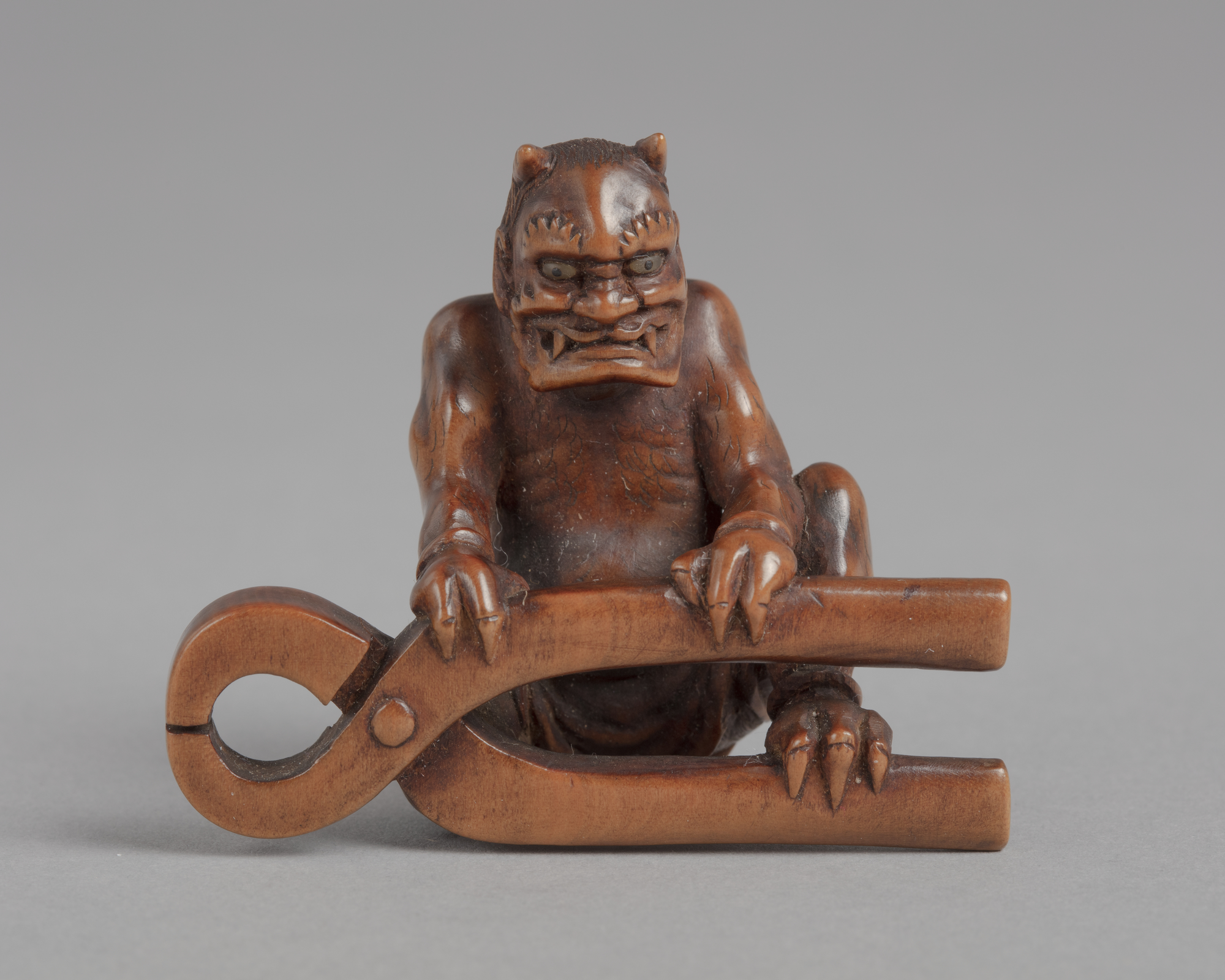 A Japanese ivory netsuke of a horned demon seated before a large pair of pliers with its hands resting on it.