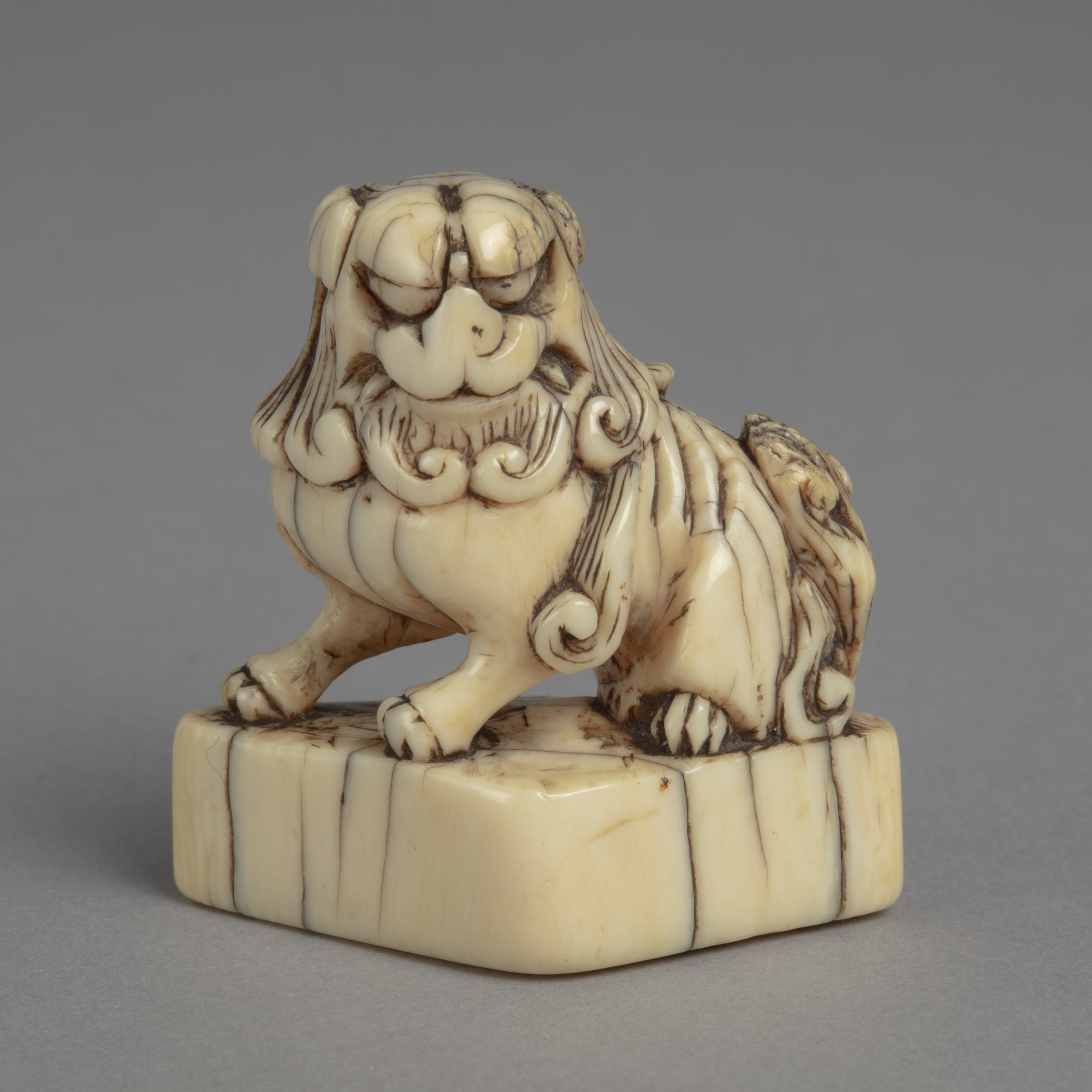 A Japanese ivory netsuke of a Chinese Buddhist lion, seated on a triangular plinth.